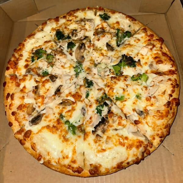 A whole Tatiana Pizza with white sauce, chicken, mushrooms, and broccoli in a cardboard box.