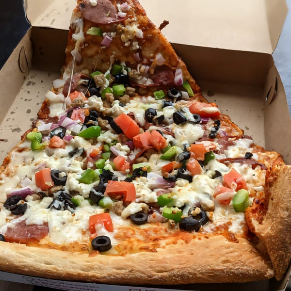 A giant slice of Tatiana Pizza with diverse toppings, including pepperoni, olives, and bell peppers, in a box.
