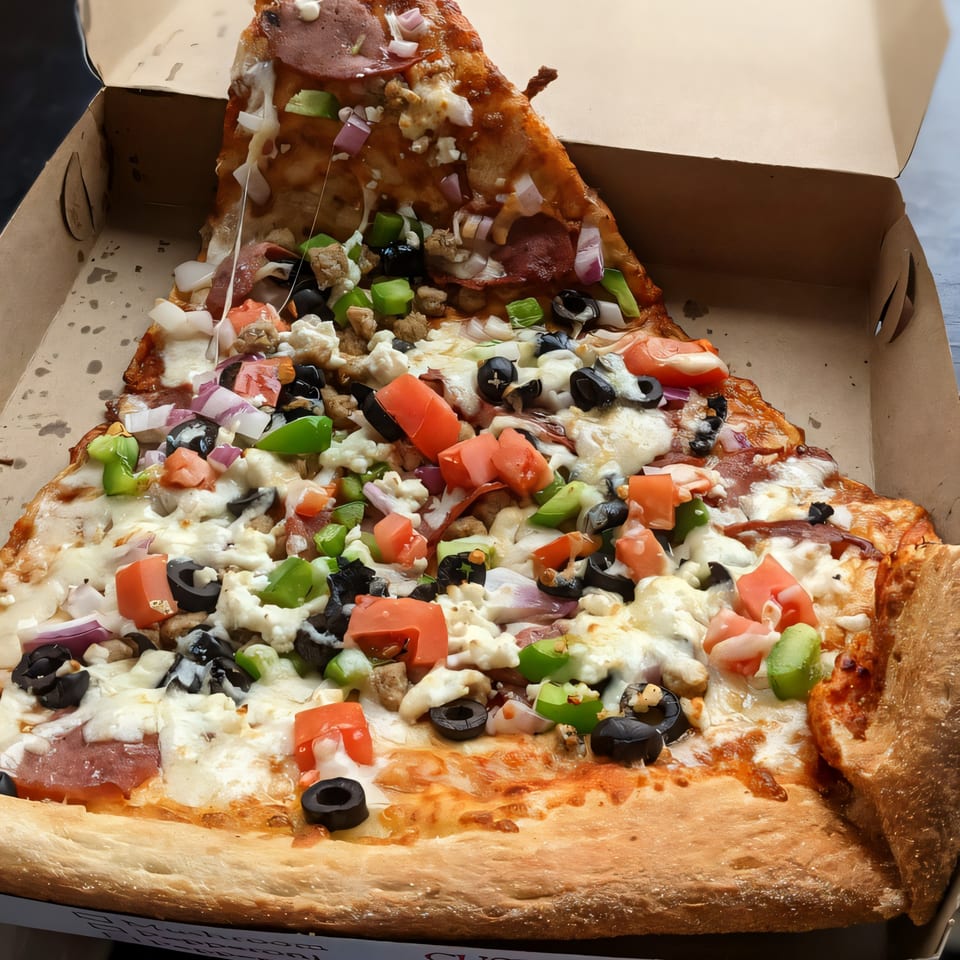 A giant slice of Tatiana Pizza with diverse toppings, including pepperoni, olives, and bell peppers, in a box.